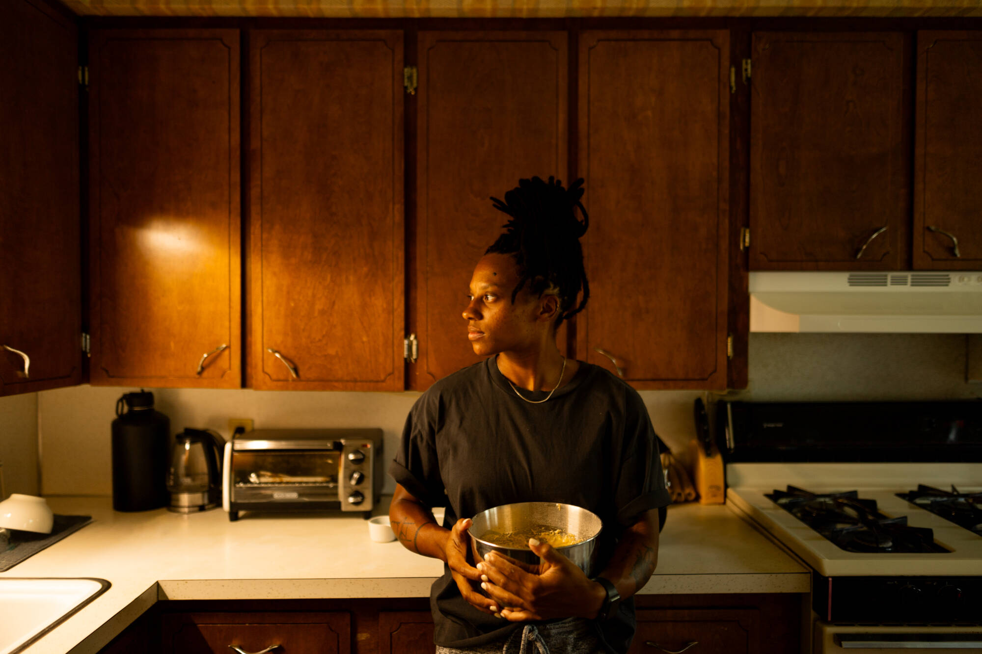 Color photograph of a person holding a metal bowl in a kitchen, looking away from the camera.